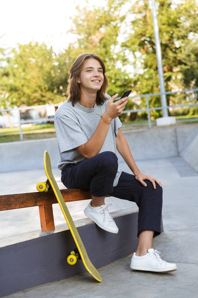 Image of cheerful young skater guy sit in the park with skateboard using mobile phone. - Фото, зображення
