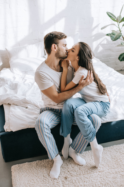 beautiful young couple in pajamas kissing while sitting on bed in morning - Foto, Imagem