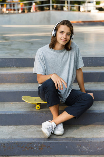Happy young teenage boy spending time at the skate park, listening to music with headphones, sitting on skateboard - Fotografie, Obrázek
