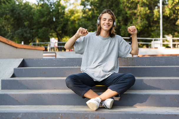 Smiling young teenage boy spending time at the skate park, listening to music with headphones, sitting on skateboard, dancing - Foto, imagen