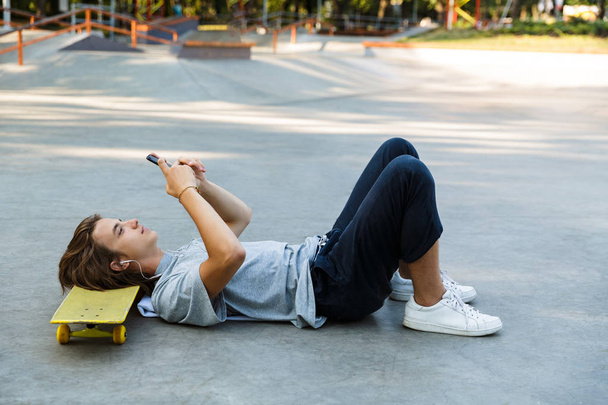 Smiling young guy spending time at the skate park, listening to music with headphones, laying on skateboard, holding mobile phone - Fotoğraf, Görsel