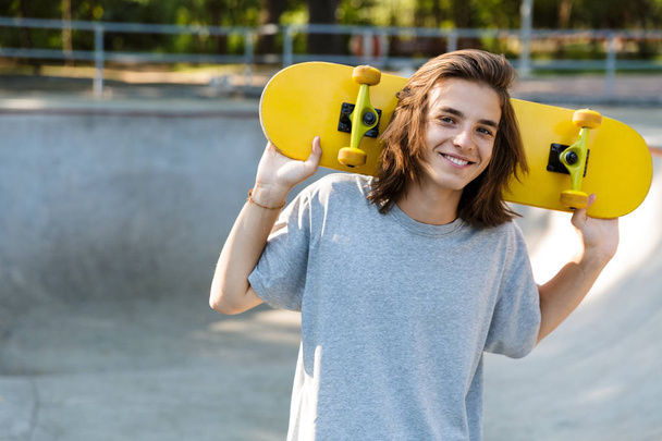 Joyful young teenge boy spending time at the skate park, holding a skateboard - Photo, Image