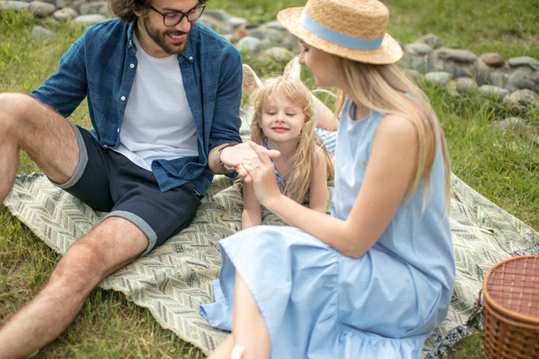 happy Family picnicking outdoors with their cute daughter, blue clothes, woman in hat - Photo, image