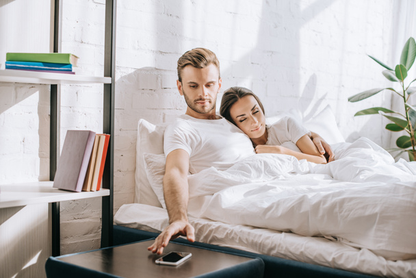 komea nuori mies rentouttava sängyssä tyttöystävän kanssa ja tavoittaa älypuhelimen
 - Valokuva, kuva