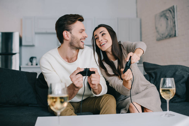 happy young couple with glasses of wine playing retro video game on couch at home - 写真・画像