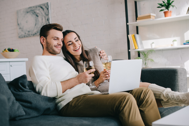 laughing young couple using laptop and drinking wine together on couch at home - Φωτογραφία, εικόνα