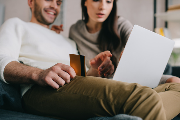 Schönes junges Paar beim E-Shopping mit goldener Kreditkarte und Laptop auf der heimischen Couch - Foto, Bild