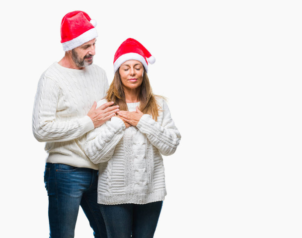 Латиноамериканская пара средних лет в рождественской шляпе на изолированном фоне улыбается руками на груди с закрытыми глазами и благодарным жестом на лице. Концепция здоровья
. - Фото, изображение