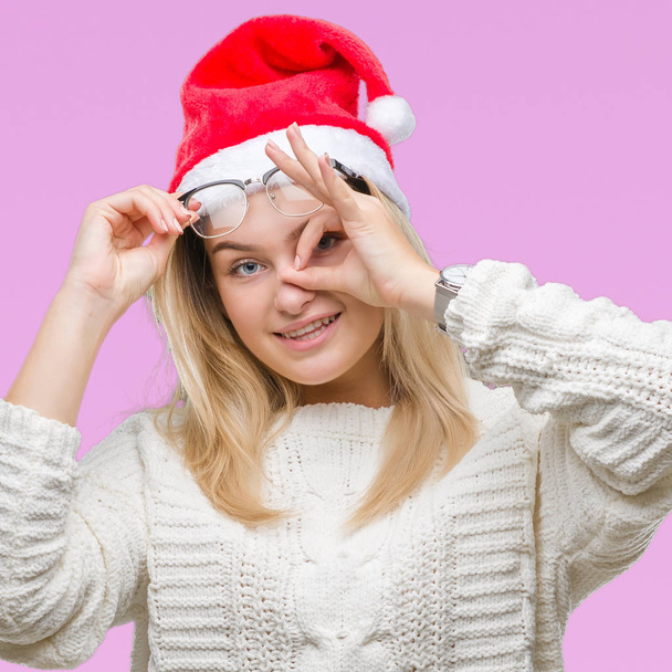 Mujer caucásica joven con sombrero de navidad sobre fondo aislado haciendo buen gesto con la mano sonriente, ojos mirando a través de los dedos con cara feliz
. - Foto, imagen