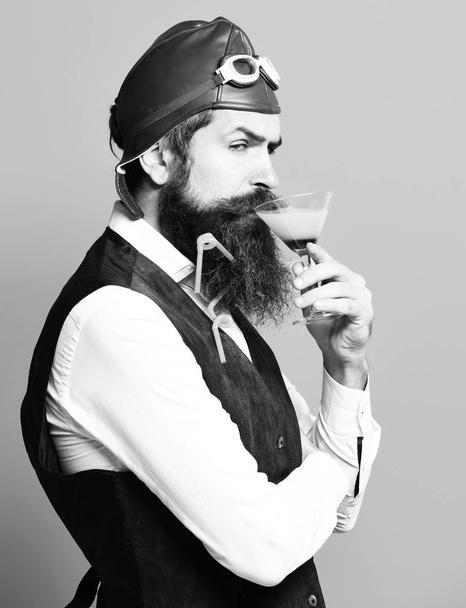 handsome bearded pilot or aviator man with long beard and mustache on serious face holding glass of alcoholic beverage in vintage suede leather waistcoat with hat and glasses on red studio background - Valokuva, kuva