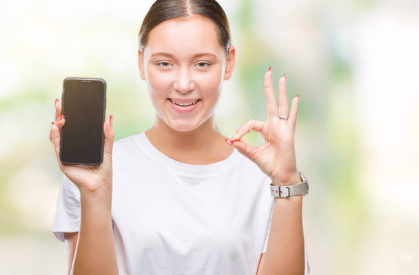 Jovem bela mulher branca mostrando tela do smartphone sobre fundo isolado fazendo sinal ok com os dedos, excelente símbolo
 - Foto, Imagem