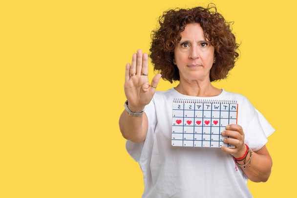 Старша жінка середнього віку, що тримає календар менструацій на ізольованому фоні з відкритою рукою робить знак зупинки з серйозним і впевненим виразом, жест захисту
 - Фото, зображення