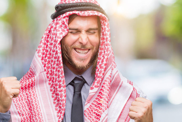 Молодой красивый арабский мужчина с длинными волосами носит keffiyeh на изолированном фоне очень счастлив и взволнован делая жест победителя с поднятыми руками, улыбаясь и крича об успехе. Концепция празднования
. - Фото, изображение