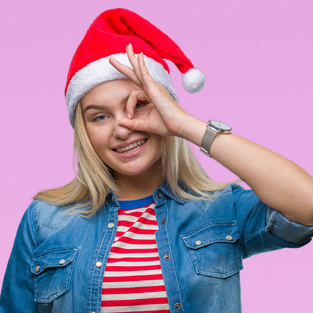 Young caucasian woman wearing christmas hat over isolated background doing ok gesture with hand smiling, eye looking through fingers with happy face. - Photo, Image