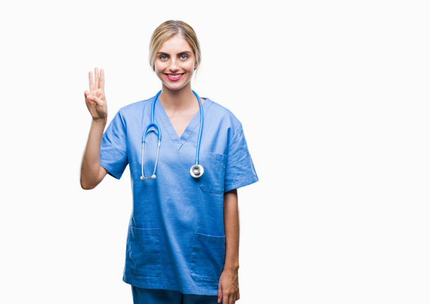 Joven hermosa doctora rubia cirujana enfermera mujer sobre fondo aislado mostrando y apuntando hacia arriba con los dedos número tres mientras sonríe confiado y feliz
. - Foto, Imagen