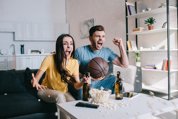 expressive young couple with beer cheering for basketball game at home - Photo, Image