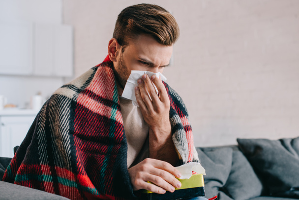 sick young man sneezing with paper napkins sitting on couch - Photo, Image