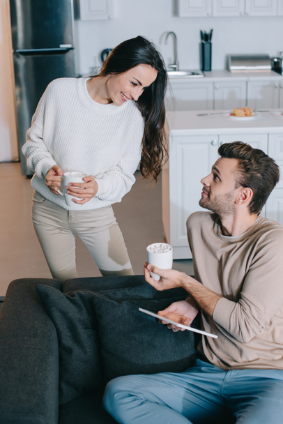 улыбающаяся молодая пара с чашками какао и таблетками, расслабляющие вместе дома
 - Фото, изображение