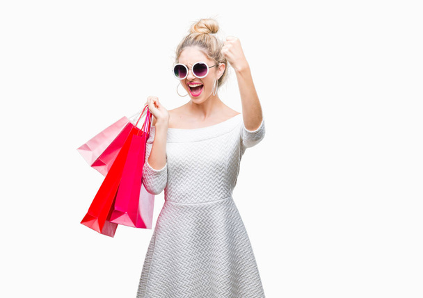 Young beautiful blonde woman holding shopping bags over isolated background annoyed and frustrated shouting with anger, crazy and yelling with raised hand, anger concept - 写真・画像