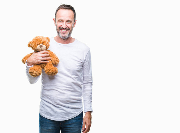 Middle age senior hoary man holding teddy bear over isolated background with a happy face standing and smiling with a confident smile showing teeth - Valokuva, kuva