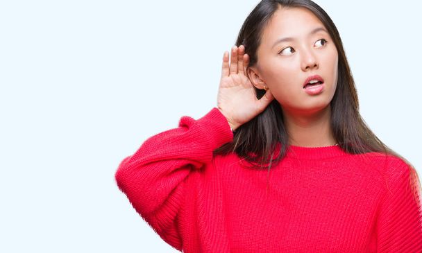 Giovane donna asiatica che indossa maglione invernale su sfondo isolato sorridendo con mano sopra l'orecchio ascoltando una voce o pettegolezzi. Concetto di sordità
. - Foto, immagini