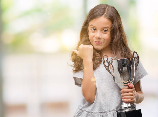 Bruna ragazza ispanica in possesso di un trofeo che indica e mostra con il pollice verso l'alto di lato con il viso felice sorridente
 - Foto, immagini
