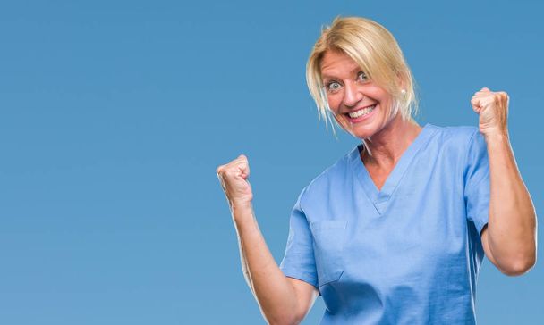 Blonde Frau mittleren Alters in Krankenschwesteruniform über isoliertem Hintergrund sehr glücklich und aufgeregt dabei Siegergeste mit erhobenen Armen, lächelnd und schreiend nach Erfolg. Festkonzept. - Foto, Bild