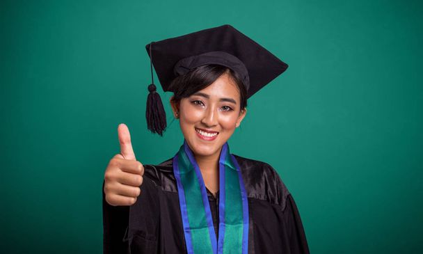 schöne lächelnde asiatische Studentin mit Abschlussmütze und Robe, die eine Daumen-hoch-Geste macht, während ihr rechter Arm in die Kamera zeigt. Nahaufnahme Porträt über grünem Banner - Foto, Bild