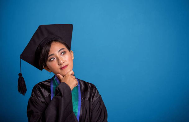 Searching for an answer. Thoughtful face expression of an Asian female student wearing graduation robe an hat, closeup portrait over blue background - Photo, Image
