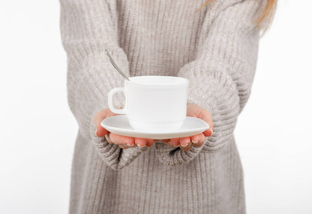 Young woman in sweater holding a white cup and saucer in hands, drinking hot coffee. - Foto, afbeelding
