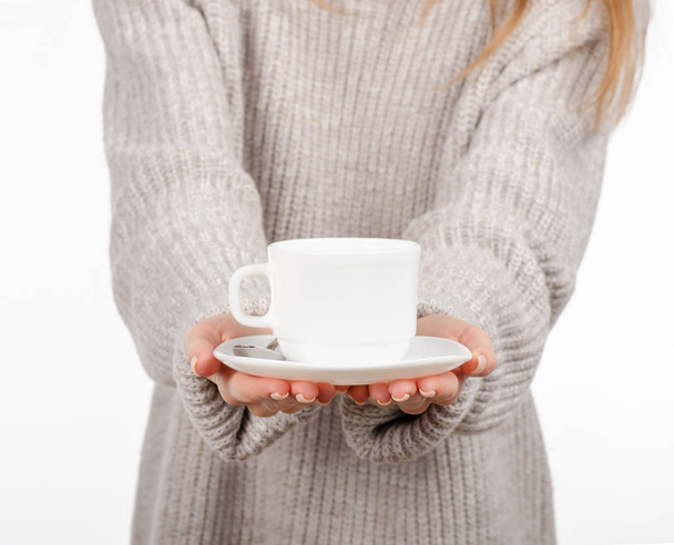 Young woman in sweater holding a white cup and saucer in hands, drinking hot coffee. - Фото, изображение