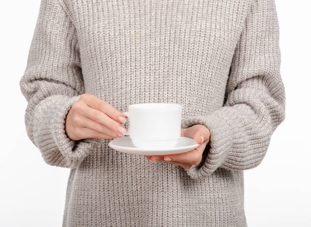 Young woman in sweater holding a white cup and saucer in hands, drinking hot coffee. - 写真・画像