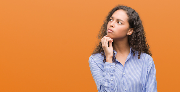 Young hispanic business woman with hand on chin thinking about question, pensive expression. Smiling with thoughtful face. Doubt concept. - Photo, Image