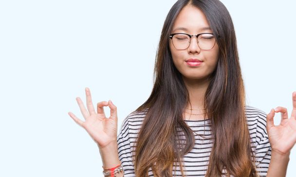 Young asian woman wearing glasses over isolated background relax and smiling with eyes closed doing meditation gesture with fingers. Yoga concept. - Fotó, kép