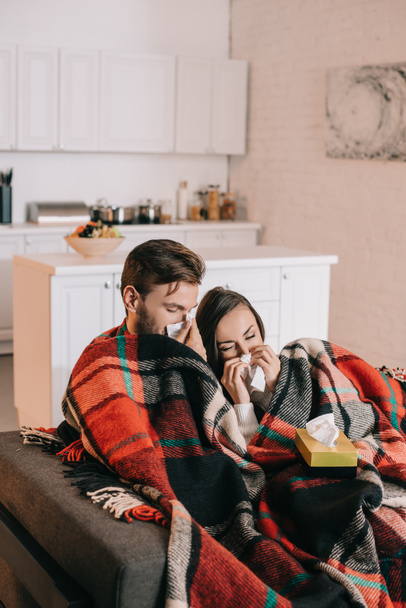 diseased young couple with paper napkins sneezing while sitting on couch under plaid at home - Foto, imagen