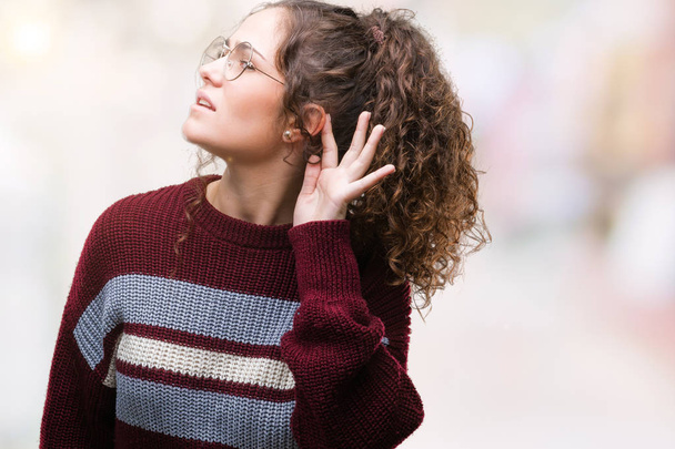 Beautiful brunette curly hair young girl wearing glasses over isolated background smiling with hand over ear listening an hearing to rumor or gossip. Deafness concept. - Photo, Image