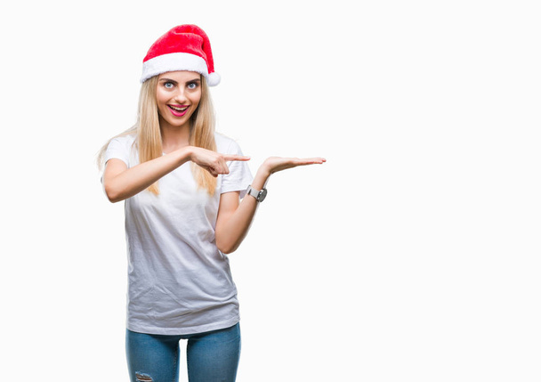 Young beautiful blonde woman christmas hat over isolated background amazed and smiling to the camera while presenting with hand and pointing with finger. - Photo, Image