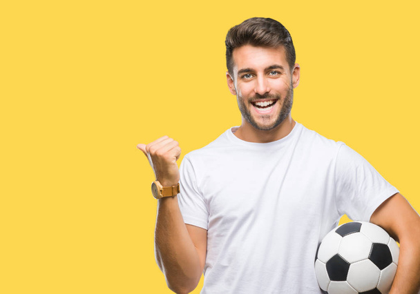 Όμορφος νεαρός, κρατώντας την μπάλα ποδοσφαίρου ποδοσφαίρου πέρα από το απομονωμένο υπόβαθρο κατάδειξης και δείχνοντας με αντίχειρα μέχρι την πλευρά με χαρούμενο πρόσωπο χαμογελαστό - Φωτογραφία, εικόνα