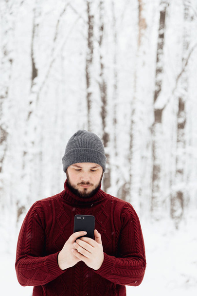 adam kırmızı örgü kazak ve kış ormanın içinde kar üzerinde duran bir telefon tutarak kot - Fotoğraf, Görsel