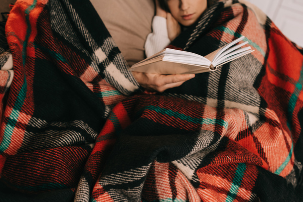 cropped shot of young couple reading book together on couch under plaid at home - Фото, изображение