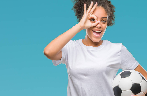 Fiatal gyönyörű afro amerikai gazdaság futball futball-labda, több mint elszigetelt háttér és boldog arc mosolyog, csinál a kéz-szem nézegette ujjak ok jele - Fotó, kép