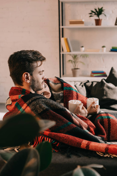 beautiful young couple with cups of cocoa with marshmallow relaxing on couch under plaid at home - Photo, Image
