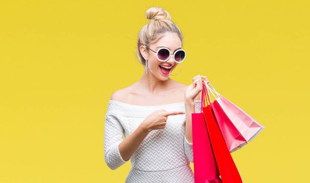 Młody piękny blond kobieta trzymając torby na zakupy na na białym tle bardzo szczęśliwy, wskazując z dłoni i palców - Zdjęcie, obraz