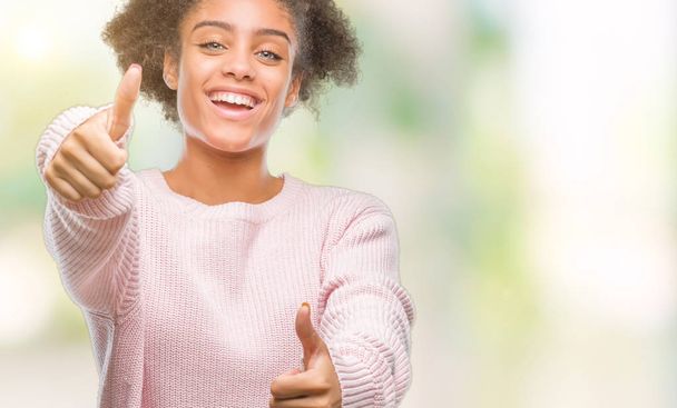 Young afro american woman wearing winter sweater over isolated background approving doing positive gesture with hand, thumbs up smiling and happy for success. Looking at the camera, winner gesture. - Fotoğraf, Görsel