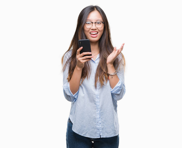 Jonge Aziatische vrouw texting met smartphone via geïsoleerd achtergrond erg blij en opgewonden, winnaar expressie vieren overwinning schreeuwen met grote glimlach en hand - Foto, afbeelding