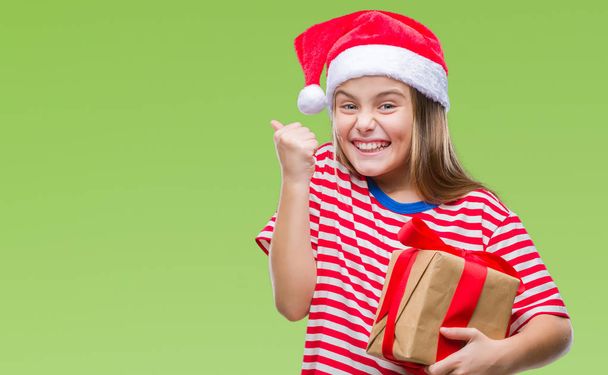 Young beautiful girl wearing christmas hat and holding gift over isolated background screaming proud and celebrating victory and success very excited, cheering emotion - Photo, Image