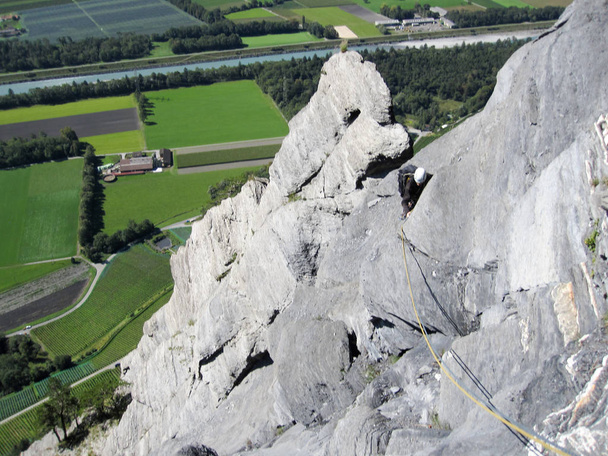 скалолаз на крутом и обнаженном известняковом скалолазании в швейцарских Альпах над пышными зелеными полями ниже
 - Фото, изображение