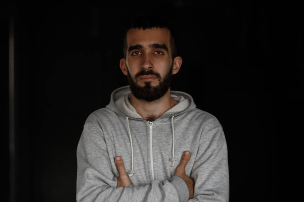 Handsome serious young man with a beard in a gray sweatshirt stands near a black wall in the studio - Photo, Image