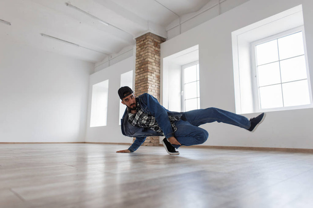 Elegante bonito jovem dançarino com um boné em um casaco de ganga azul e jeans dançando em um estúdio de dança
 - Foto, Imagem