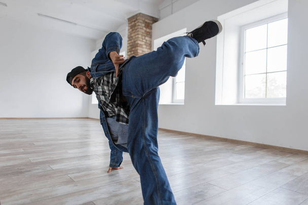 Handsome young stylish man with a cap in a denim jacket and jeans with sneakers dancing in a dance studio - 写真・画像